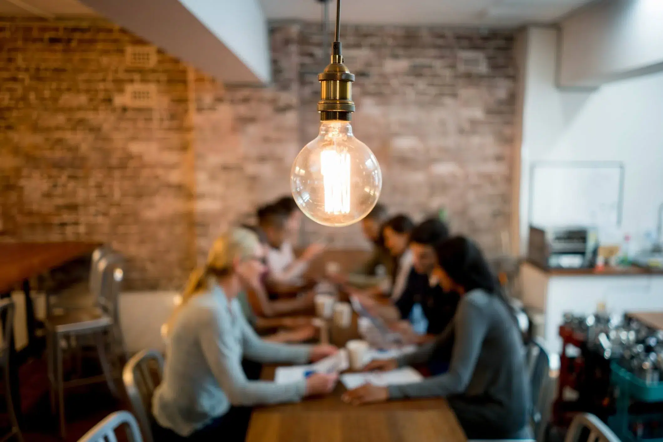 A Light Bulb Above A Table Of People Signifying Good Ideas Happen When We Work Together